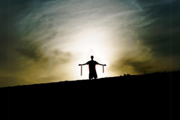 A silhouette of a person standing with arms outstretched, holding broken chains, against a dramatic sky with a glowing light behind them, symbolizing liberation and freedom.